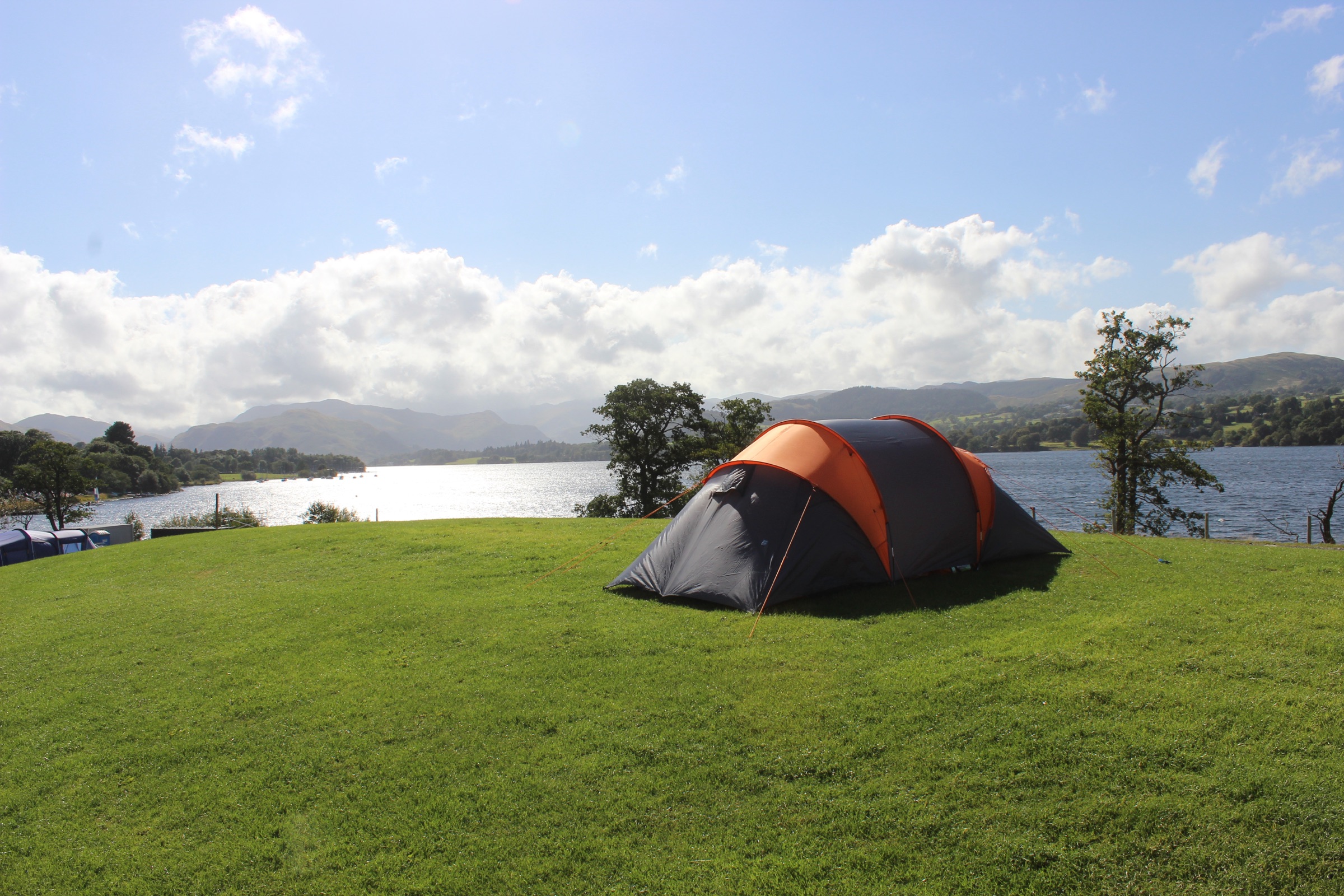 An orange and grey tent on a grassy rise above a sunlit lake, mountains in the distance